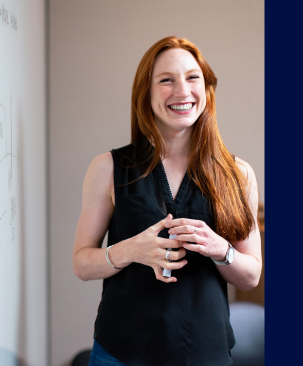 A cheerful woman with red hair holds a whiteboard, exuding warmth and enthusiasm through her smile.