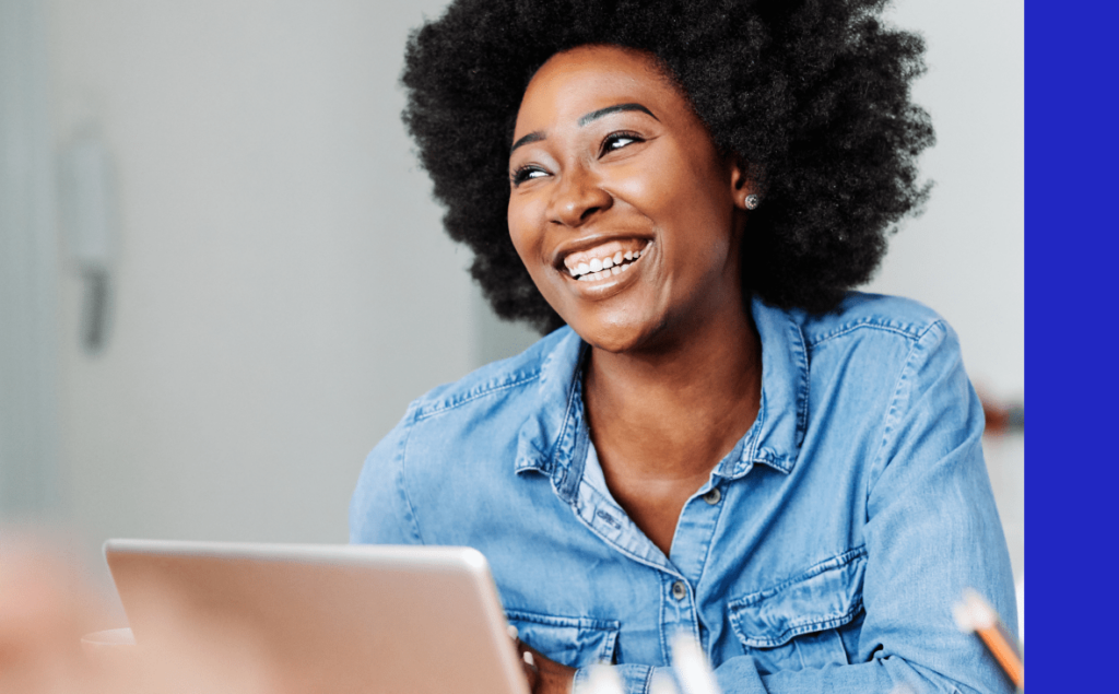 A woman with afro hair smiles brightly while working on her laptop, exuding joy and engagement in her task.